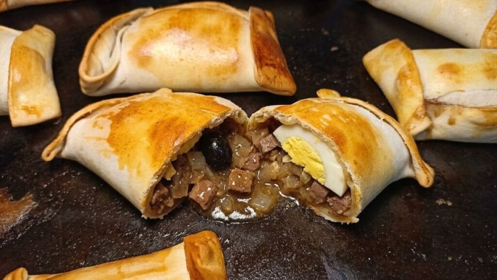 Lata de horno con empanadas chilenas doradas recién horneadas, destacando al centro una empanada abierta que muestra un pino jugoso con picado de carne, cebolla, huevo duro y aceituna negra.